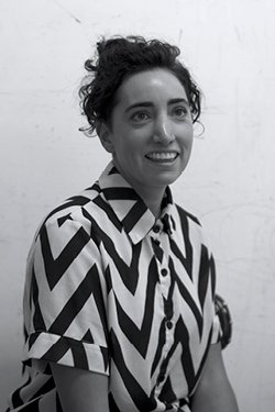 Black and white portrait of woman looking slightly off centre, with curly black hair tied up, and wearing a black and white zig zag patterned button up shirt.