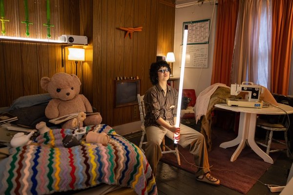 Artist EJ Son wearing sunglasses and sitting on a chair in a room with a wood-panelled wall, holding an LED light tube erect. Objects around the room include a sewing machine on a table and a bed with a blanket, teddy bear and pots strewn across it.