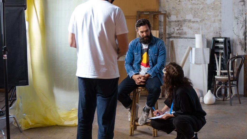 Three people in an artist studio. One has their back to the frame, he is wearing white tshirt and jeans. One is sitting on a bar stool, he is wearing a tshirt with the Aboriginal flag and looking down at another person crouched on the floor writing notes.