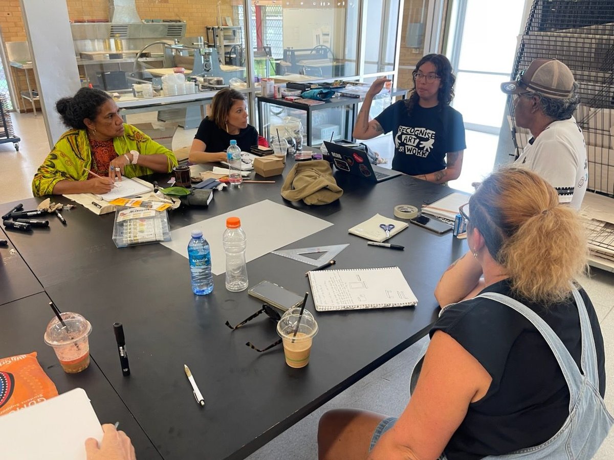 Five people sit around a dark table in an art studio, with notebooks, laptops, and drinks spread across it, as Georgia Mokak leads a discussion in their black "Recognise Art as Work" t-shirt.