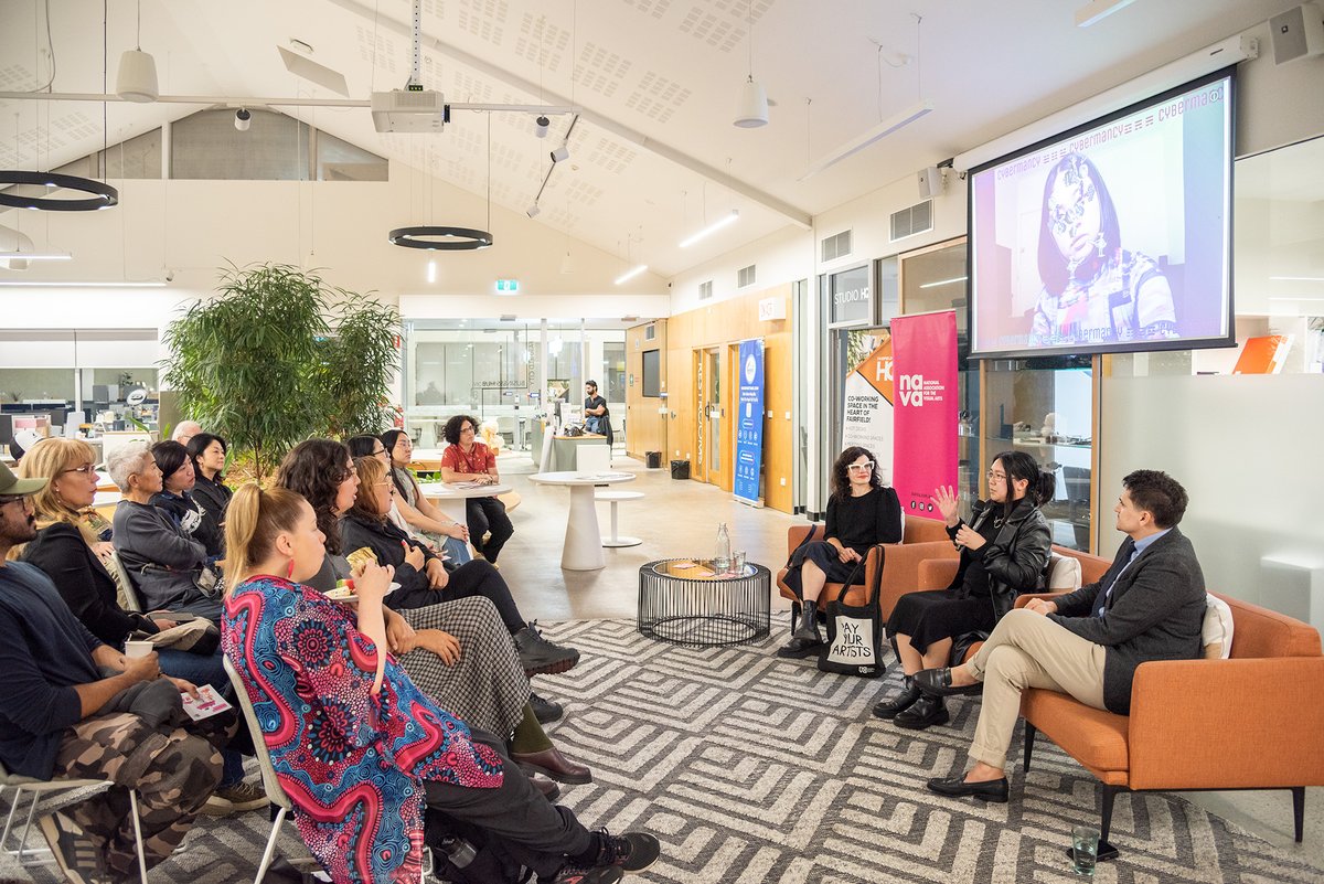 Photo of three people sitting in orange armchairs on right with projector screen above them and an audience sitting in rows on left. 