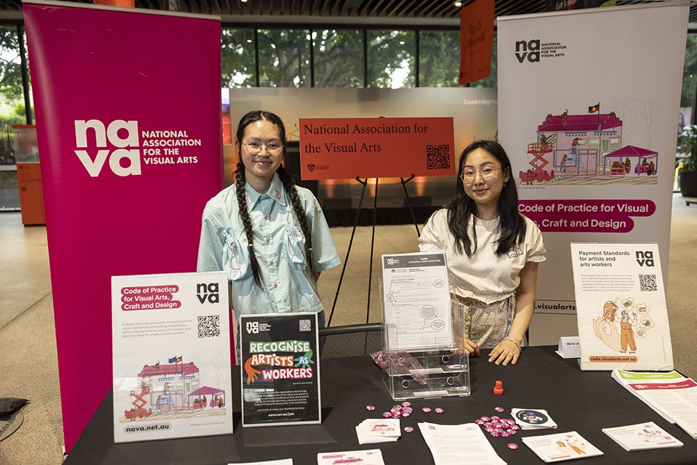 Two people smile at a NAVA booth displaying banners, posters and pamphlets promoting the Code of Practice for Visual Arts, Craft and Design.