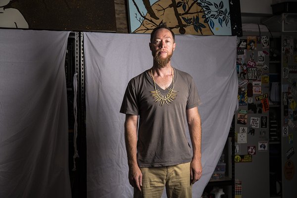 Image of a man wearing a grey v-neck t-shirt and beige/khaki pants. He is wearing a straw-coloured necklace with long pointy sticks attached one by one along the bottom.