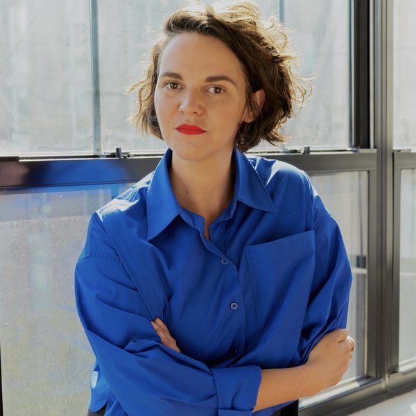 Liz is leaning with her arms crossed against a window sill, wearing a bright blue shirt with black pants. She has shoulder-length wavy brown hair and is wearing red lipstick.