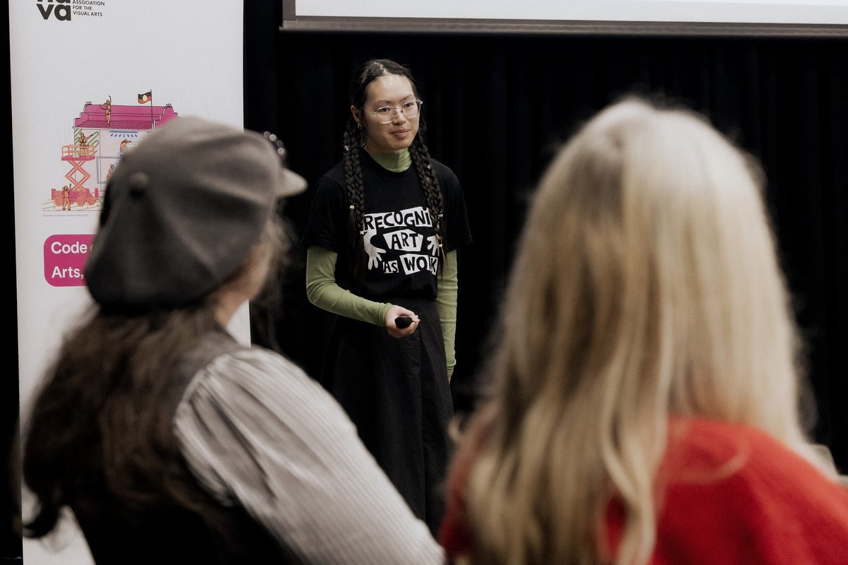 Photo of person presenting at a NAVA event wearing a “Recognise Art as Work” t-shirt, with audience members listening.