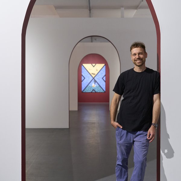 Photo of Ryan standing at a series of archways, an out of focus painting in the background. He is wearing a grey shirt and smiling at the camera.
