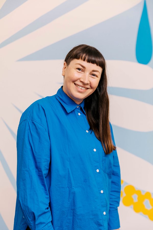 Artist April Phillips smiles at the camera with their long brown hair pushed to the front on one side. April is wearing a bright blue long sleeve shirt with white buttons.