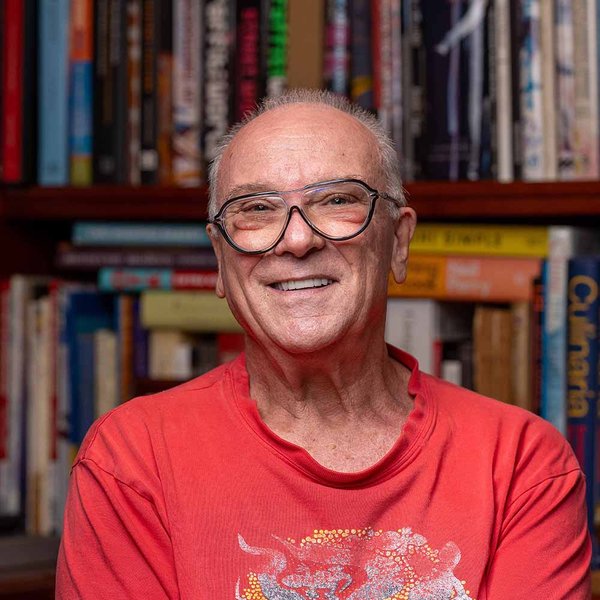 Portrait of Brian Tucker. He is wearing a red t-shirt, thin framed glasses and sitting in front of a large bookcase.