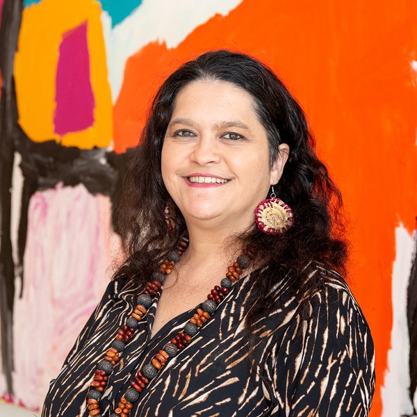 Head and shoulders portrait of Bianca Beetson, she is smiling, has dark brown wavy hair, is wearing a dark patterned blouse and standing in front of colourful painting of bright orange, black, white, pink and yellow.