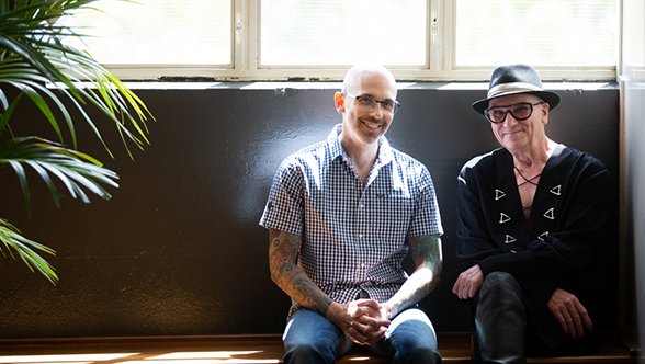 Image of two men sitting beside each other, in front of a black wall indoors. 