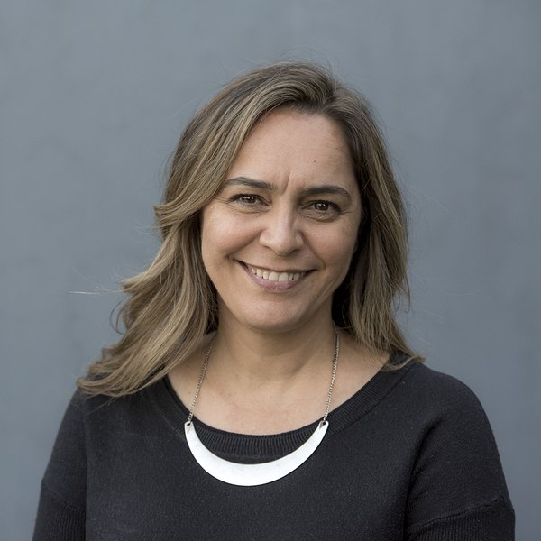 Portrait of Clothilde Bullen on grey background. She is smiling, has long wavy brown hair, and is wearing a black top and silver necklace. 