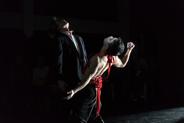 Photograph of two dances facing each other with their heads tilted back, wearing red and black with harsh stage lighting. The background is black.