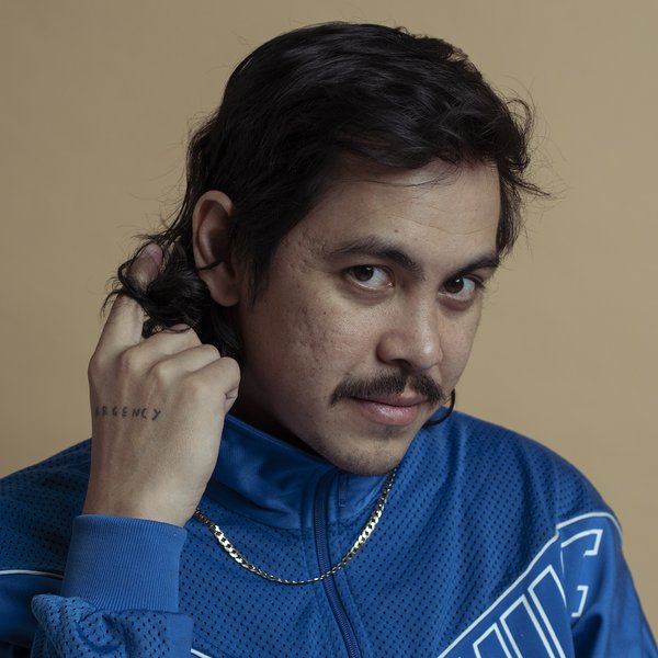 Head and shoulders portrait of Abdul Abdullah. His head is tilted and he is looking at the camera. He has dark brown hair and moustache, is wearing electric blue Nike jacket and a gold chain. The word 'urgency' tattooed on his hand which is near his face.