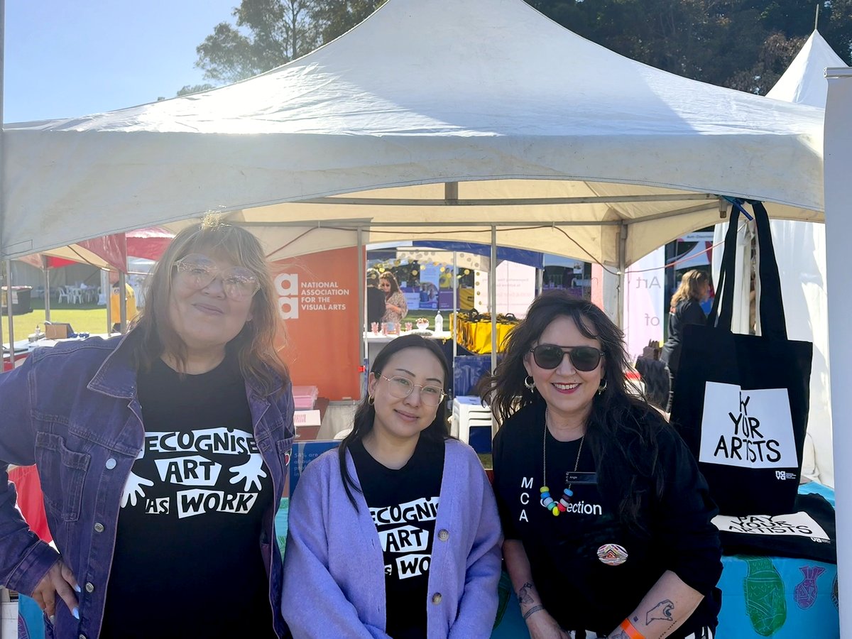 Three people wearing black t-shirts stand at the NAVA stall at a festival, with NAVA signage and ‘Pay Your Artists’ tote bags visible behind them.
