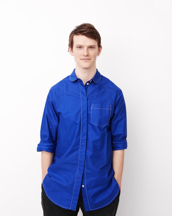 Photo of Dylan standing in front of a white background, he is wearing a blue shirt, has his hands in his pockets and is smiling at the camera.