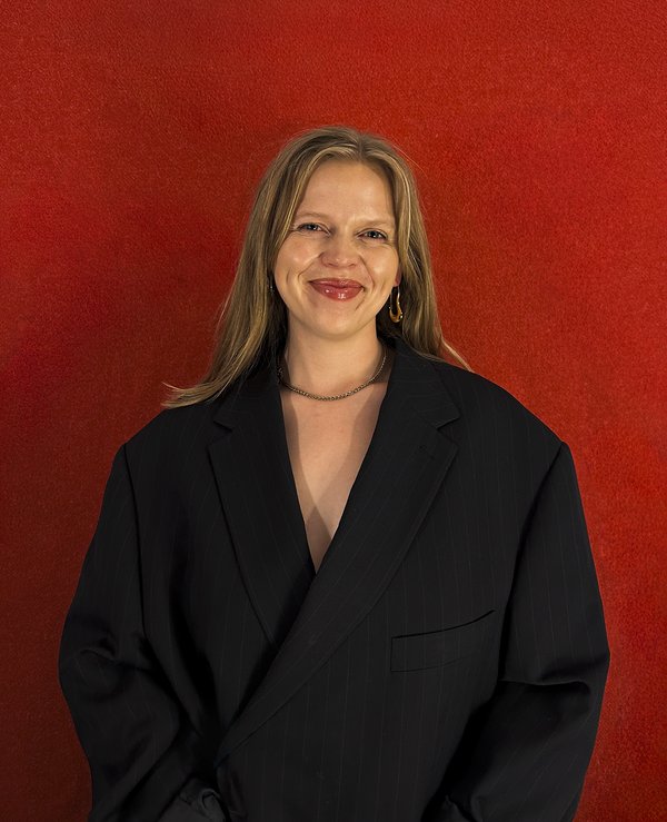 Photo of Georgia, standing against a red background, she wears a black coat, has long hair and is smiling at the camera.