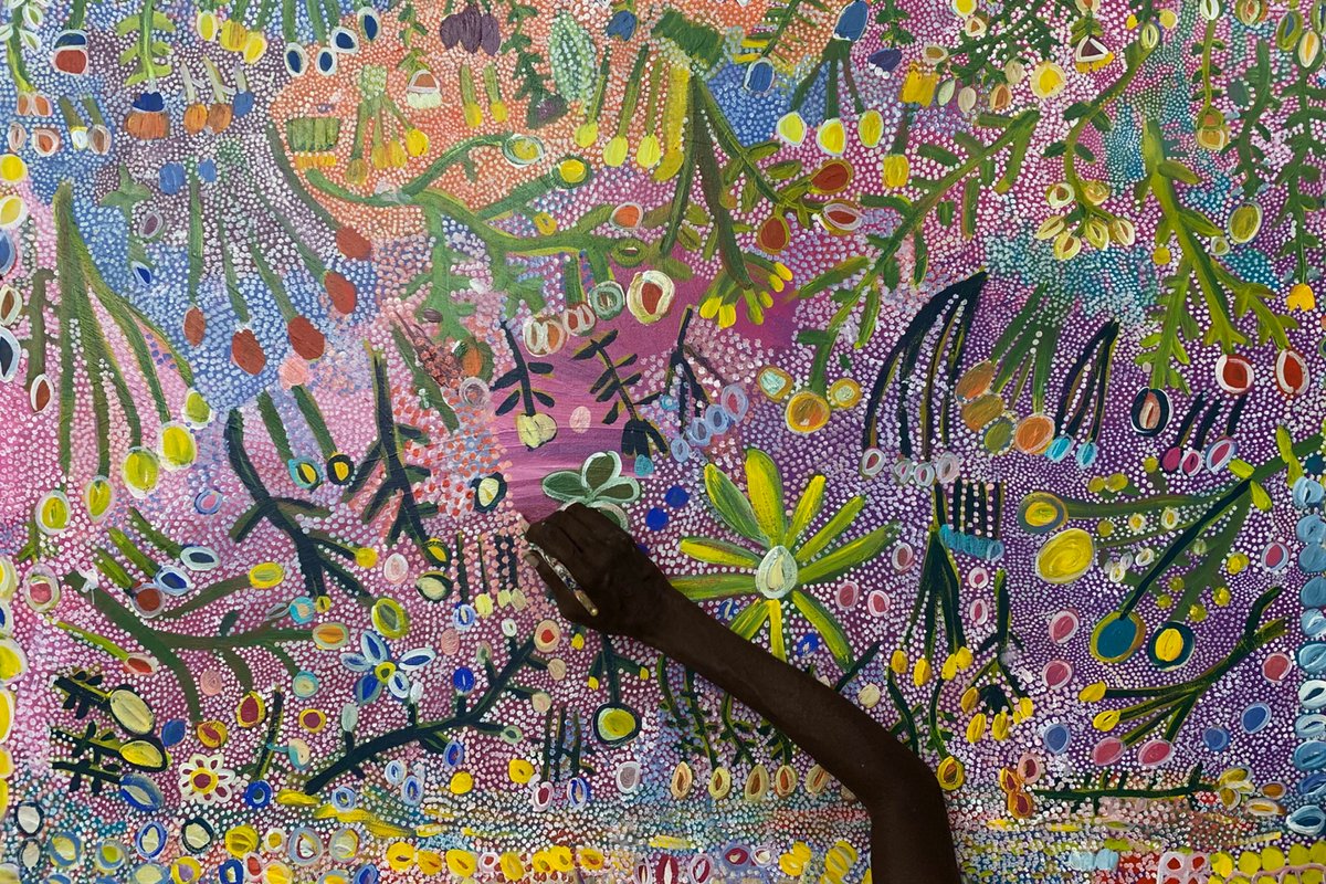 Aerial view of a large painting of flowers and plants with a purple background. A hand is holding a brush and adding to detail to the centre of the work.