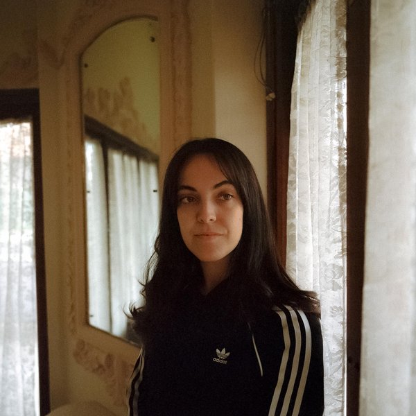 Image of a woman from the midriff up. She has long black hair and is wearing a black adidas jacket with three white stripes down her sleeves. She is looking to the right. Behind her are a mirror and two windows with white lace curtains.