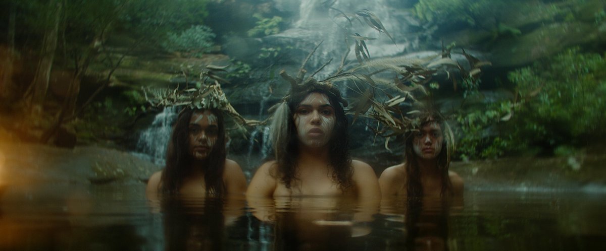 An image of three people immersed up to their chests in water. They have white markings on their faces and look directly into the camera. Behind them are trees, mossy rocks and a waterfall. The light of the image is soft and hazy.