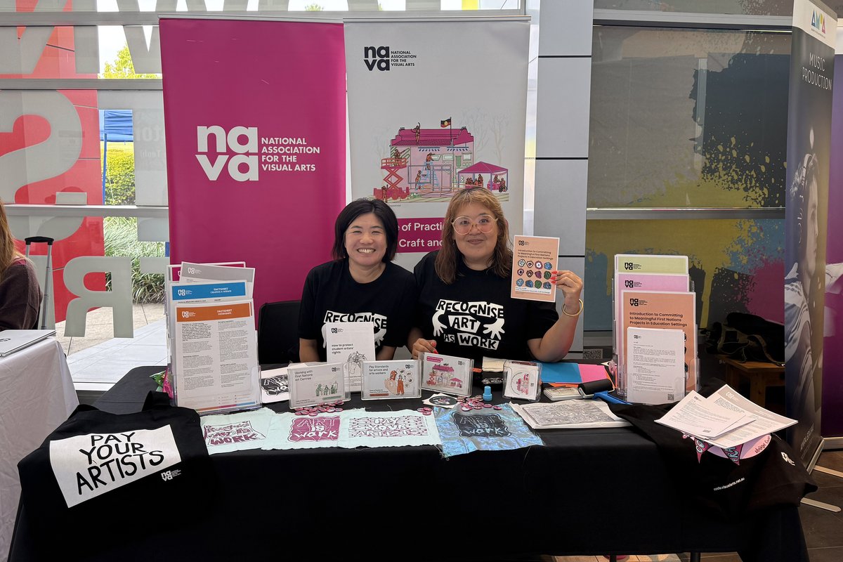 Two people wearing "Recognise Art as Work" T-shirts sit behind a NAVA information stall displaying publications, campaign materials and a "Pay Your Artists" sign. NAVA banners are visible behind them.