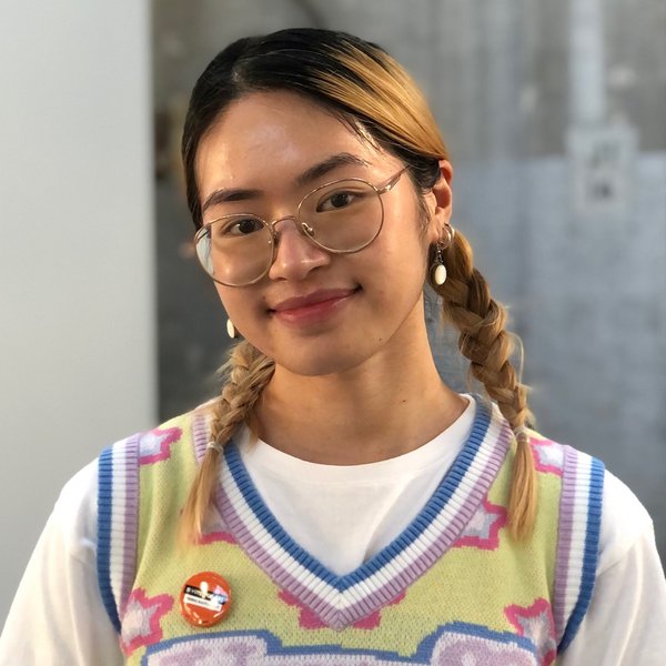 Head and shoulders portrait of Emma Pham. She is wearing a white shirt with a colourful vest, glasses, and has her blonde hair in braids.