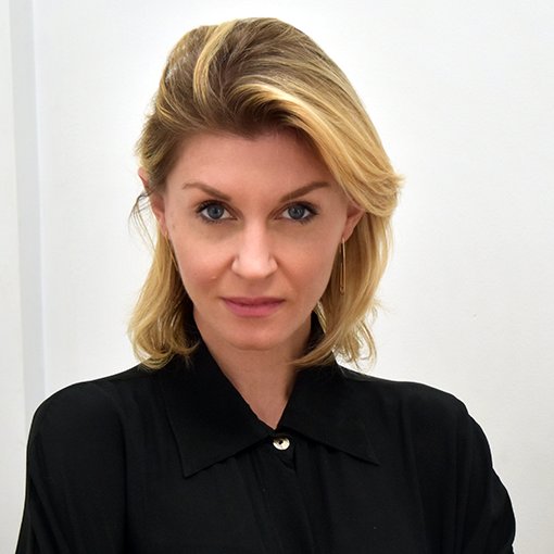 Head and shoulders portrait of Imogen Beynon. She is wearing a black long sleeve shirt, has shoulder length blonde hair and is looking at the camera. She stands in front of a white background.