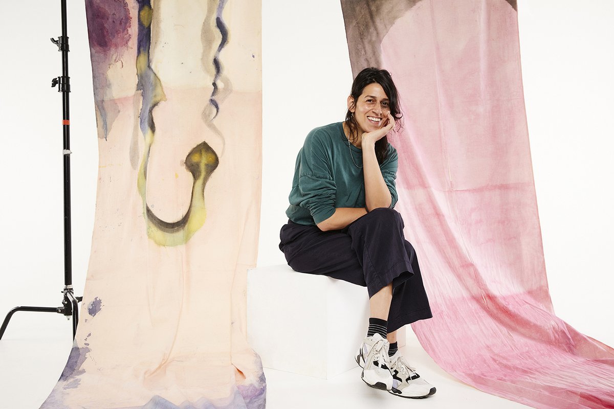 A photo of a person sitting on a white plinth, elbow leaning on her knee, her legs are crossed. She is wearing a green top, black pants and white sneakers. She has brown hair and is smiling. Ink paintings on fabric are draped in the background.