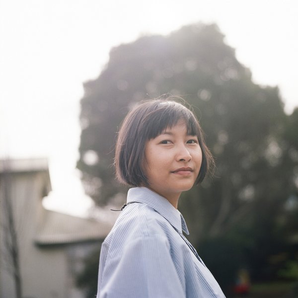 Jamie is looking slightly past the camera with their head turned. She has short, dark hair and is wearing a light blue, collared shirt. They are outdoors, with a softly blurred background featuring a large tree and a building.