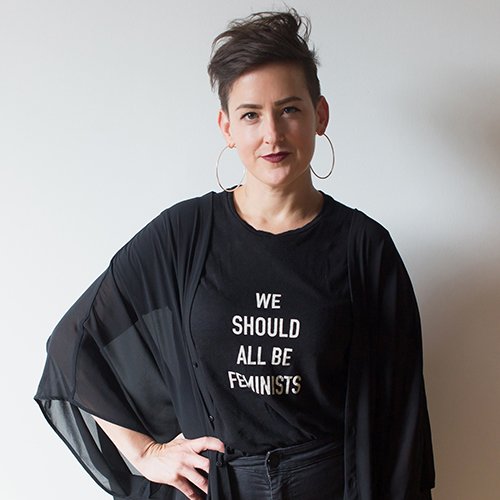 Portrait of Kate Just, she is wearing a black shirt which says 'we should all be feminists' in white text, her hand is on her hip and she is looking at the camera
