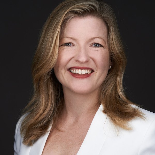 Head and shoulders portrait of Mimi Crowe. She is wearing a white jacket, has long wavy blonde hair and is smiling.