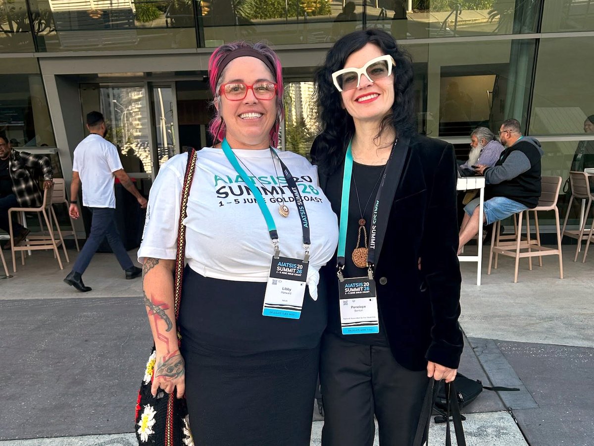Libby Harward and Penelope Benton stand smiling side by side outside the AIATSIS Summit 2026 venue, both wearing conference lanyards and name badges