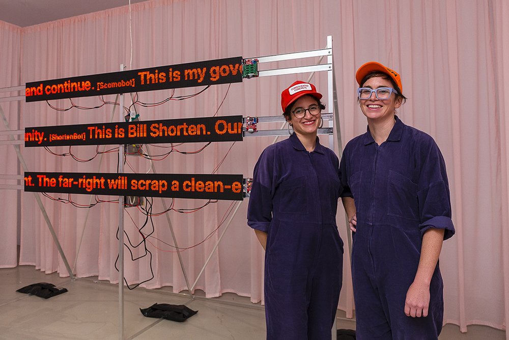 Portrait of the two artists wearing matching blue boiler suits and orange caps, standing in front of their work - a pink curtain and three LED scrolling text displays