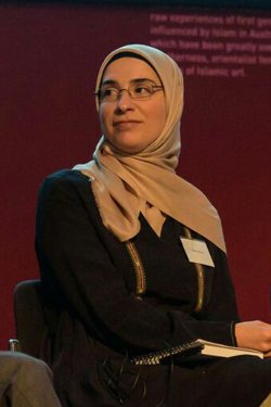 Woman wearing a black dress, clear glasses and a beige headscarf looking slightly over her shoulder. On stage speaking at a panel.