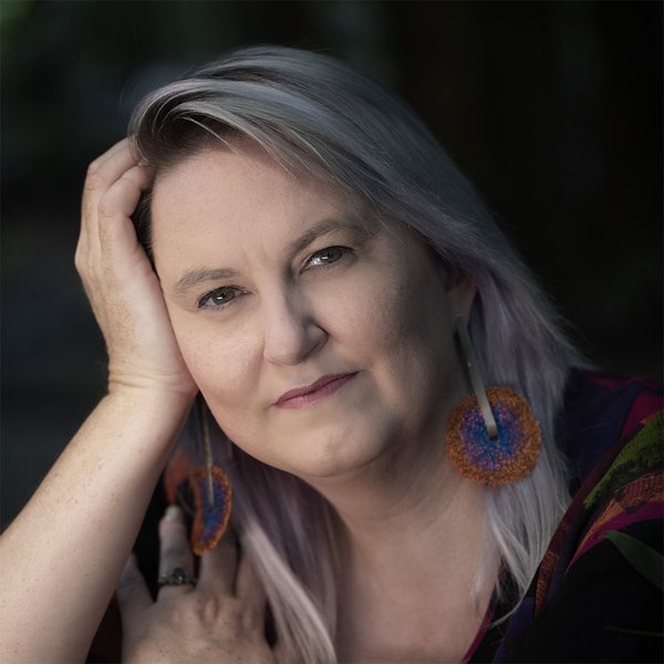 Head and shoulders portrait. Michelle is leaning her head on her hand, the other hand is on her shoulder. She has shoulder length white hair and is wearing earrings with orange and blue textile circles