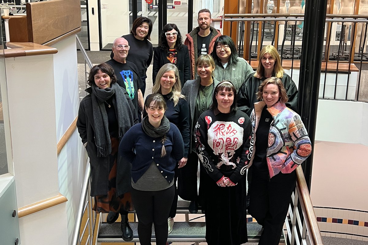 A group of thirteen people wearing warm clothing including scarves and coats stand on a stairwell indoors, posing for a group photo.