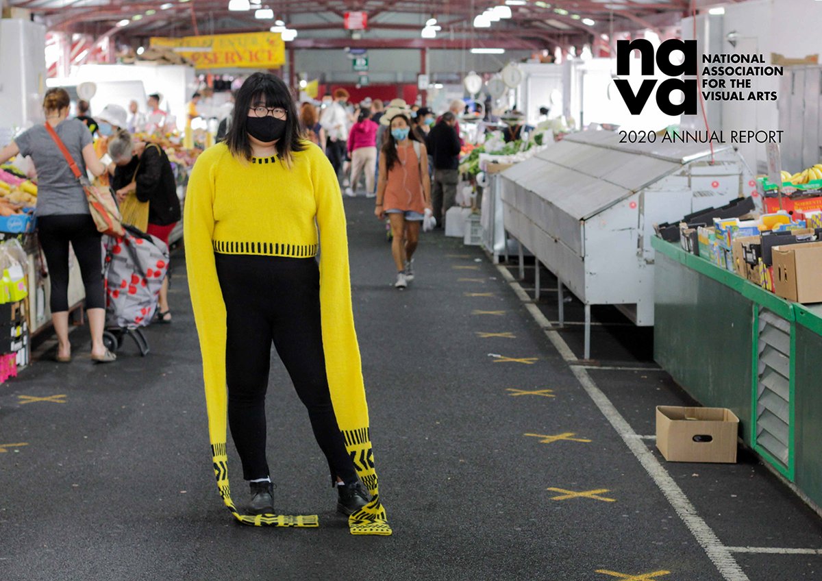 A woman is standing slightly left of centre in the aisle of a fresh produce market. She is wearing a black face-mask and a bright yellow sweater with long sleeves all the way to the ground, caution tape pattern is knitted into the lower sleeve.