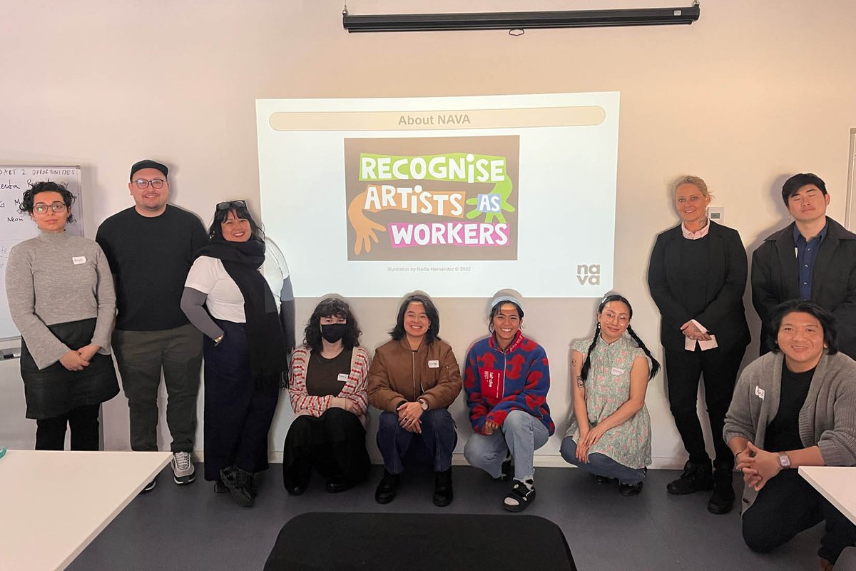 A group of ten people pose together in a room, standing and kneeling in front of a presentation slide with Nadia Fernandez’s artwork that reads, “Recognise Artists as Workers” in bold, colourful text. 
