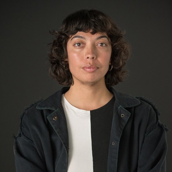 Portrait of Georgia Mokak. They have short, curly dark hair, looking directly at the camera with a neutral expression. They are wearing a black-and-white top and a dark jacket with frayed edges. The background is plain and dark.