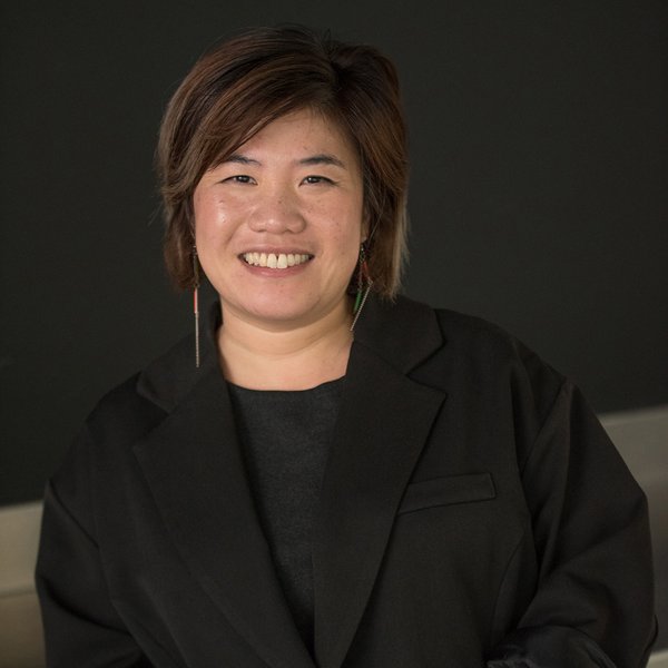 Portrait of Janel Yau wearing a black blazer, long earrings, with dark background. She has short dark brown hair with light brown highlights. 