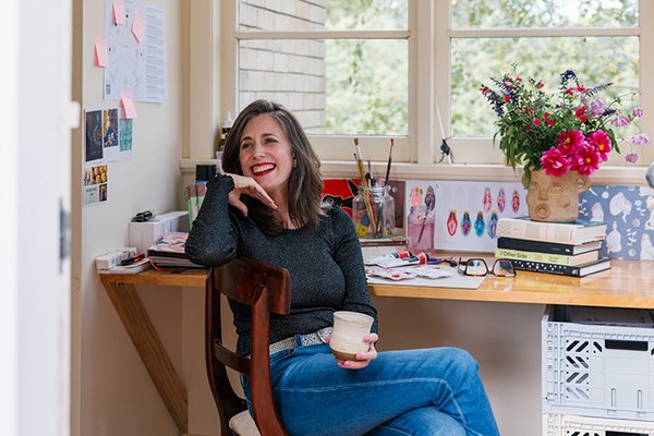 A woman smiling in her art studio, sitting in a chair and holding a tea mug. 