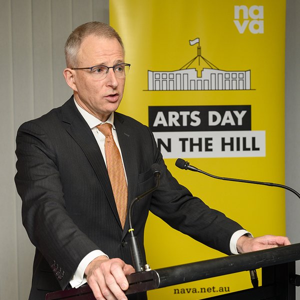 A man wearing a suit and thin framed glasses is facing the right side of the frame, his two hands are on either side of a lectern. A yellow banner in the background says Arts Day on the Hill.