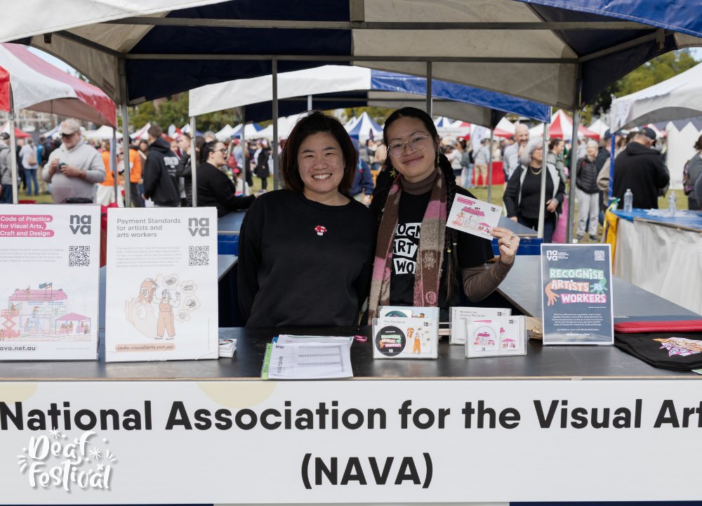 Two people smile at a NAVA booth at an outdoor festival. The booth displays posters and pamphlets promoting fair pay and recognition for artists, with a crowd visible in the background.