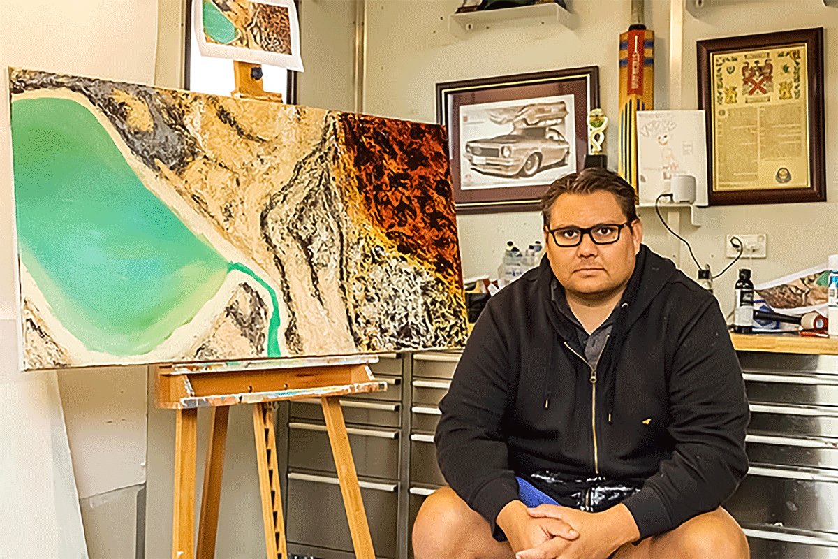 Photo of man with short black hair wearing black hoodie, black glasses and shorts, seated to the right of a work in progress painting on a wooden easel in an art studio.