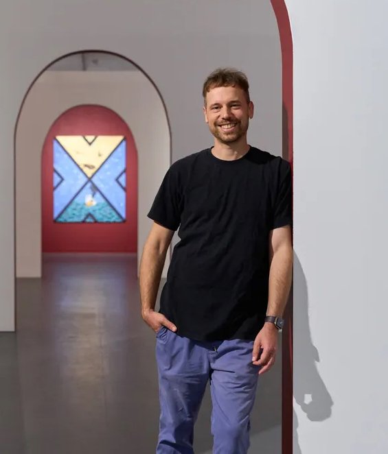 Photo of Ryan standing at a series of archways, an out of focus painting in the background. He is wearing a grey shirt and smiling at the camera.