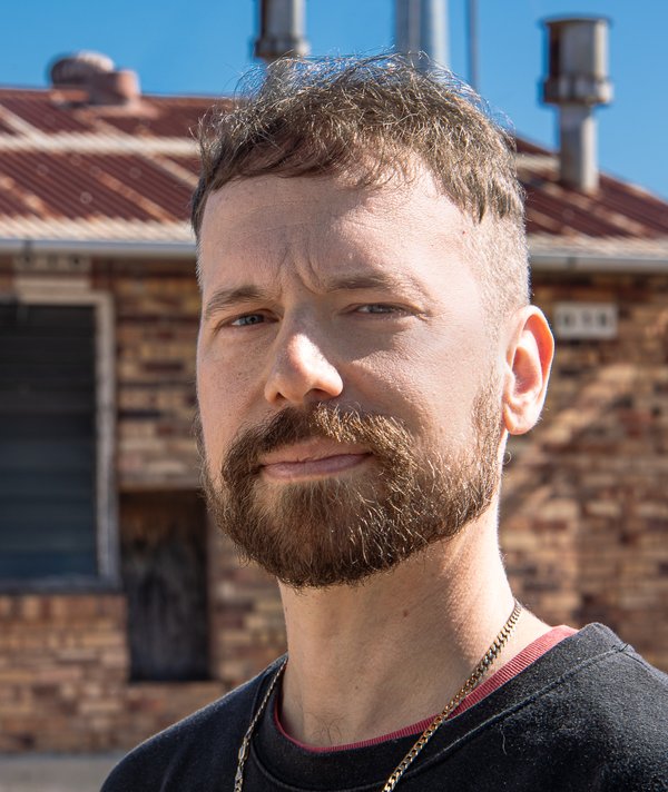 Artist Ryan Presley looks to the camera, squinting with a slight smile. He is wearing a gold chain, dark top. A rusted roof and brick building is in the background.