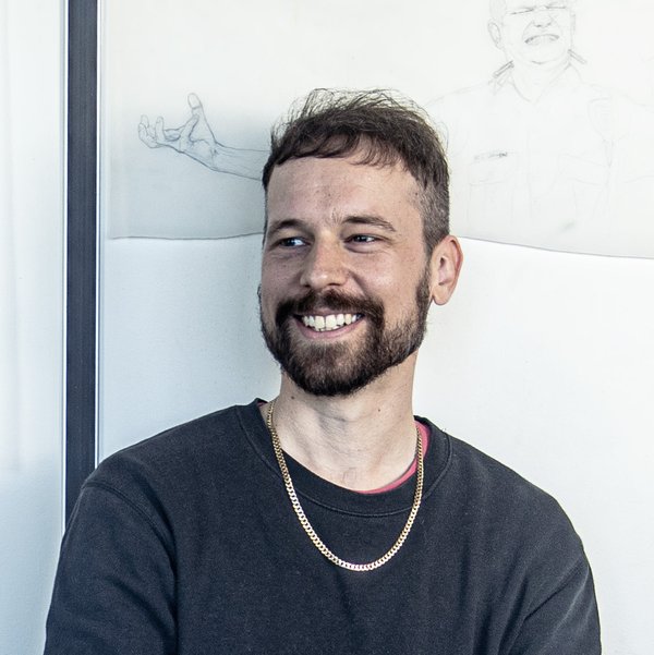 Head and shoulders portrait of Ryan Presley. He has short brown hair and a beard, and is looking out of frame and smiling. He is wearing a black jumper and gold chain. A faint pencil drawing on white paper is posted on the wall behind him.