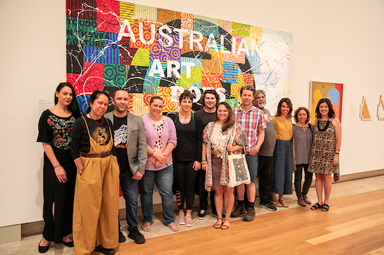 Twelve people standing in a row in front of bright painting by Richard Bell with large text that says Aboriginal Art Does. The last line is covered by the group.
