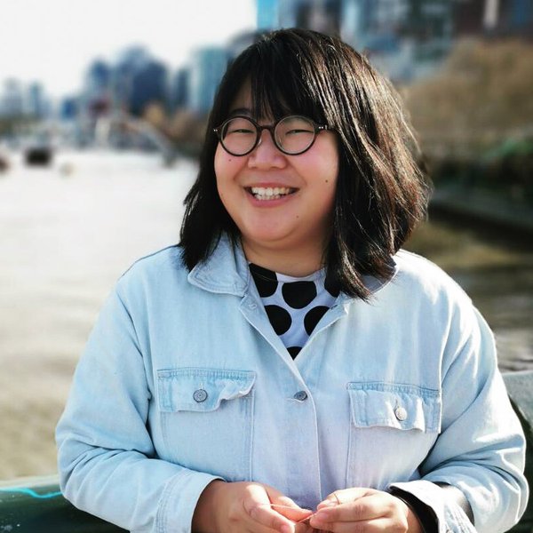 Portrait of Sophia Cai. She has dark shoulder length hair with a fringe and black round framed glasses. She is smiling and wearing a pale blue collared shirt over a black and white spot tshirt. In the background there is water and a distant cityscape. 