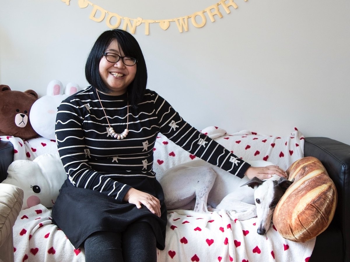 Photo of woman with shoulder length straight black hair and fringe, wearing striped black and white long-sleeve shirt, black skirt, black framed glasses and white beaded necklace sitting on a couch with one hand resting gently on her white greyhound.