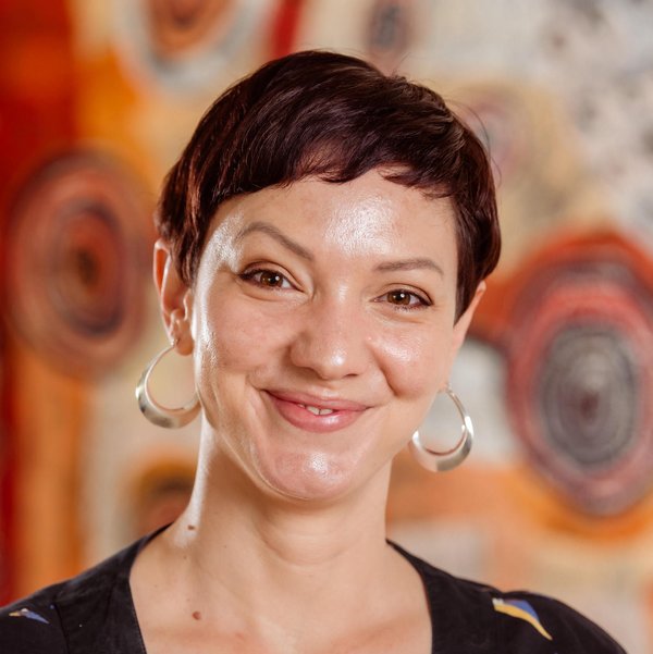 Head shot of Sophia Sambono, she has short brown hair, her head is tilted and she is smiling. She is wearing a black top and silver hooped earings. In the background is a painting of orange and red tones.